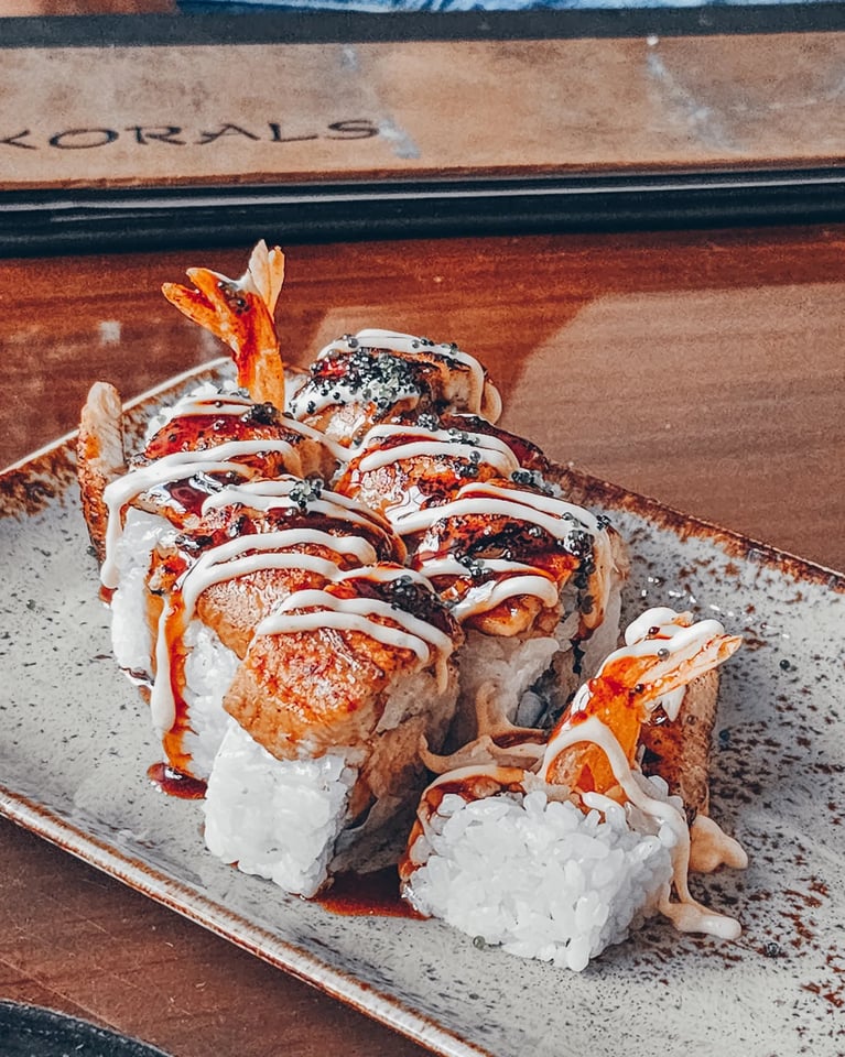 Tempura shrimp sushi roll drizzled with eel sauce, spicy mayo and sesame seeds on a ceramic plate at Korals Sushi & Cocktail Lobby Bar