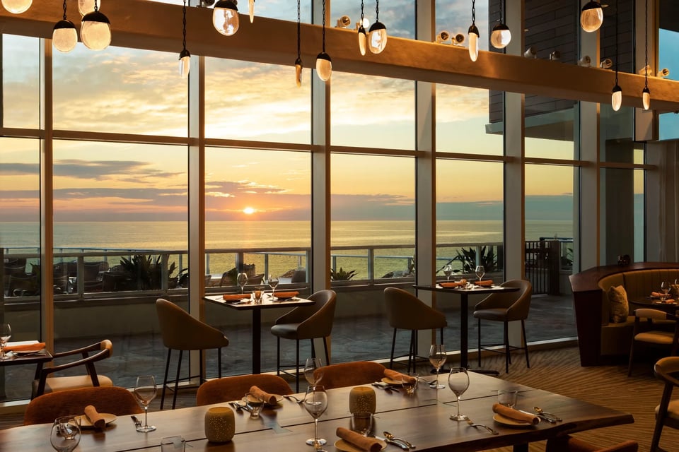 Floor-to-ceiling windows at Tesoro frame a golden Gulf sunset above set tables and pendant lights