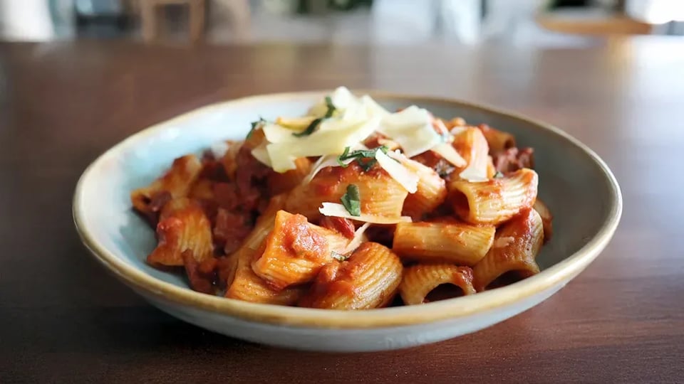 Bowl of rigatoni in tomato sauce topped with shaved cheese and basil on a wooden table