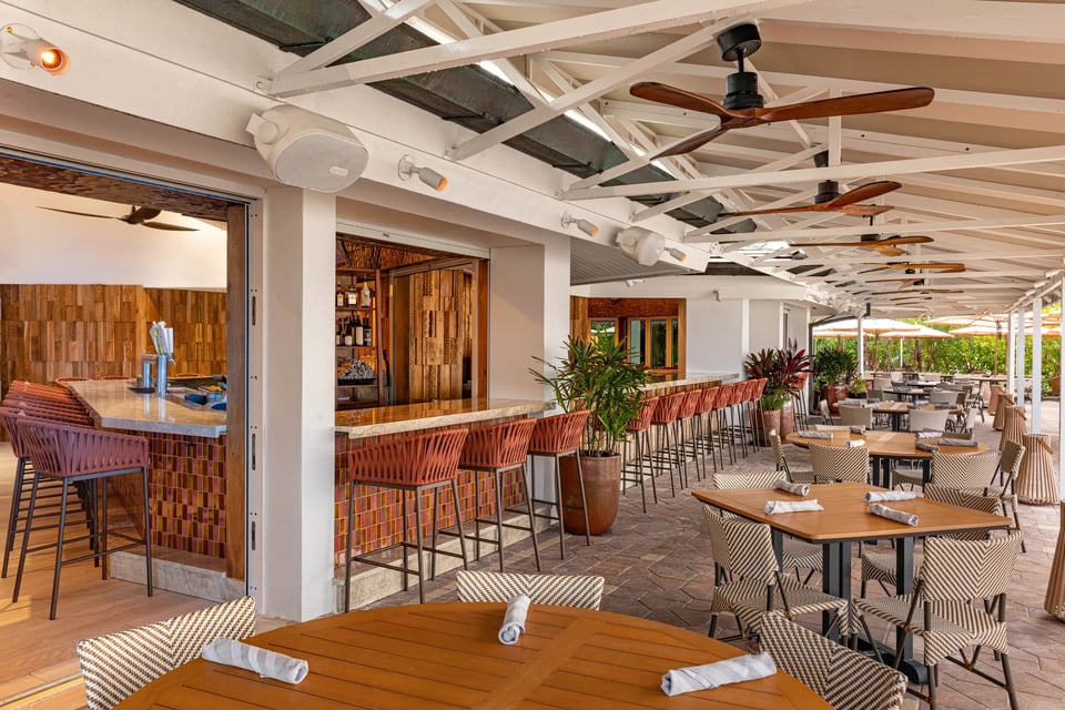 Covered open-air patio at Quinn's with woven bar stools, ceiling fans, potted palms and set wooden tables