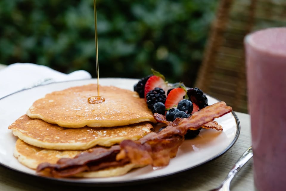 Syrup drizzling onto a stack of pancakes plated with crisp bacon and fresh berries at an outdoor table