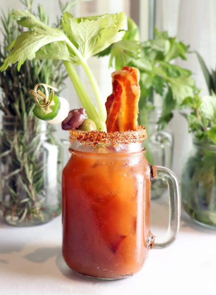 Bloody Mary in a handled mason jar with spiced rim, celery stalk, crisp bacon and skewered olives