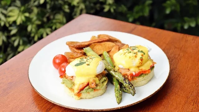 Eggs Benedict with hollandaise, asparagus, potato wedges and cherry tomatoes on a white plate at a sunlit outdoor wooden table
