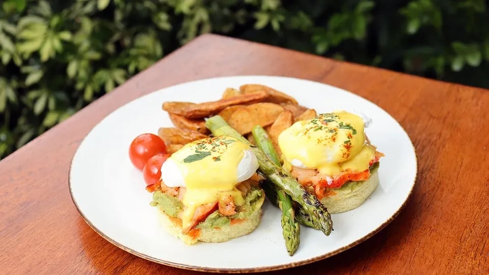 Eggs Benedict with hollandaise, asparagus, potato wedges and cherry tomatoes on a white plate at a sunlit outdoor wooden table
