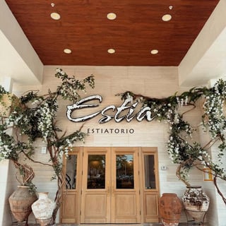Entrance to Estia Estiatorio with wooden doors, white blossom branches and large clay urns under a wood ceiling
