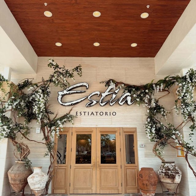 Entrance to Estia Estiatorio with wooden doors, white blossom branches and large clay urns under a wood ceiling
