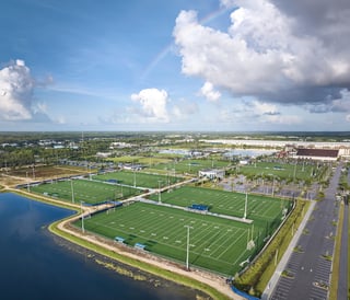 Aerial view of Paradise Coast Sports Complex showing green turf fields, a lake and a rainbow in cloudy skies