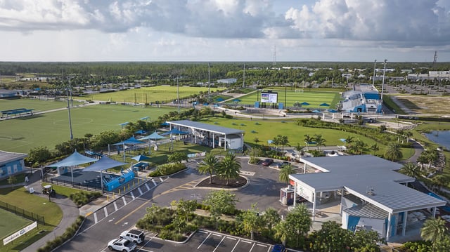 Aerial view of Paradise Coast Sports Complex with green playing fields, shade canopies, pavilions and parking lots under cloudy skies