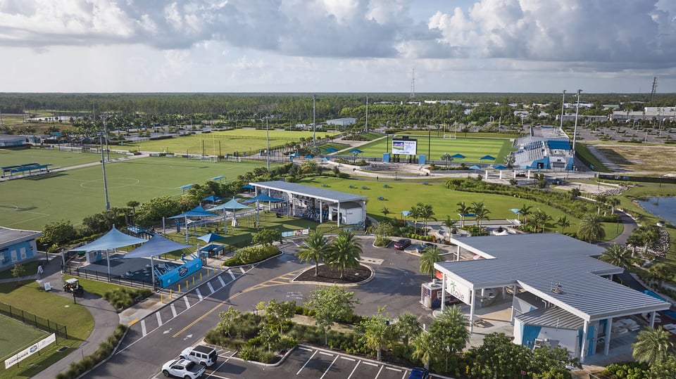 Aerial view of Paradise Coast Sports Complex with green playing fields, shade canopies, pavilions and parking lots under cloudy skies