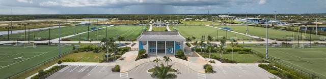Aerial panorama of Paradise Coast Sports Complex showing green playing fields, palms and the covered stadium entrance