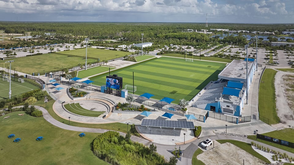 Aerial view of Paradise Coast Sports Complex showing green playing fields, blue shaded seating and a large video screen
