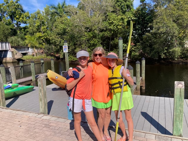 A woman and two girls in bright life vests pose with a yellow paddle on a wooden dock beside kayaks and a mangrove-lined waterway