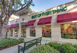 Storefront of Best of Everything with pink awnings for accessories, gifts and jewelry, shaded by trees and a green bench