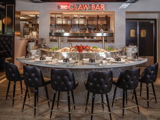 The Claw Bar's curved marble raw bar lined with black tufted stools beneath a glowing neon sign and iced seafood display