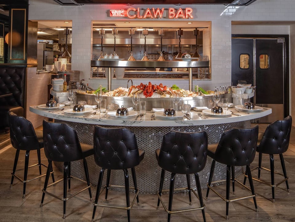 The Claw Bar's curved marble raw bar lined with black tufted stools beneath a glowing neon sign and iced seafood display