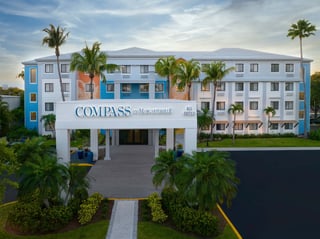 Exterior entrance of Compass Hotel by Margaritaville Naples at dusk, framed by palms under a pastel sky