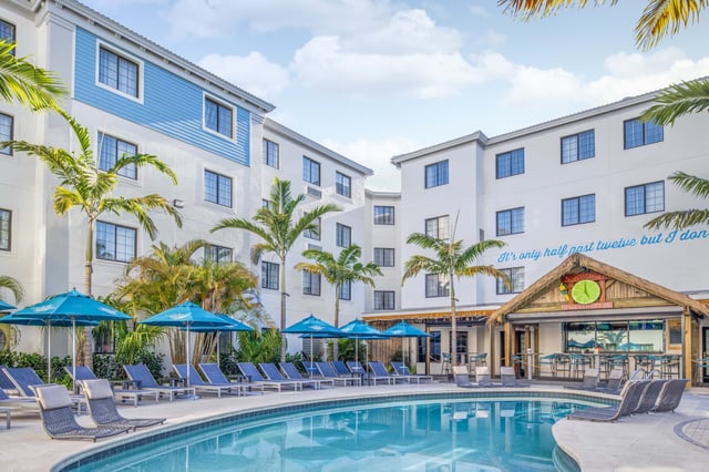 Courtyard pool with blue umbrellas and lounge chairs at Compass Hotel by Margaritaville Naples, palms and poolside bar beyond