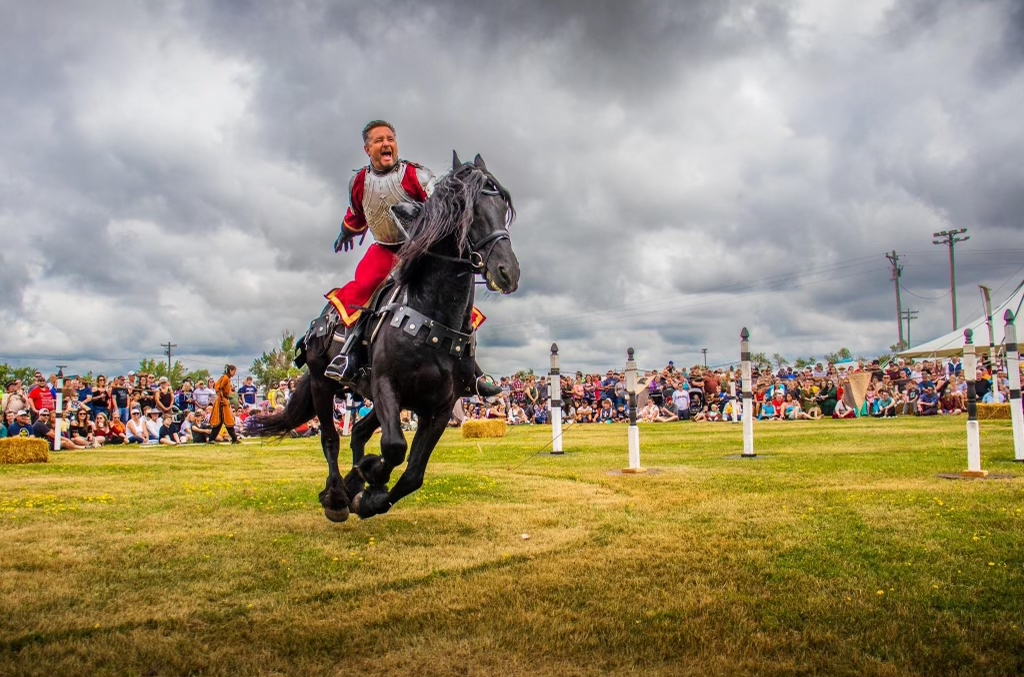 A costumed rider in red and armor gallops a black horse across a grassy field before a seated crowd under storm clouds