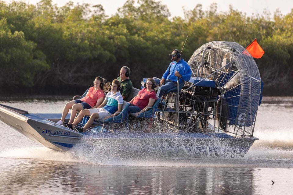 Passengers wearing headsets ride an airboat spraying water as it speeds past mangroves on an Everglades City tour