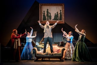 Actor standing on a sofa with arms raised as four costumed performers gesture toward him on a lamplit stage