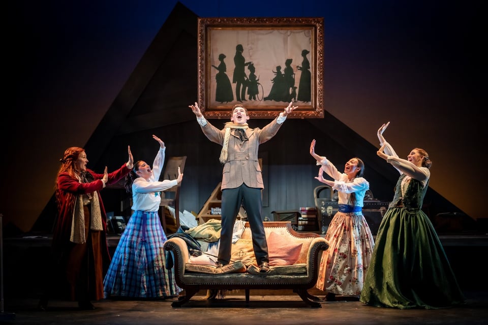 Actor standing on a sofa with arms raised as four costumed performers gesture toward him on a lamplit stage
