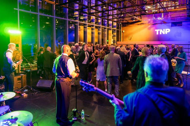 A live band performs onstage in green and blue light as a formally dressed crowd mingles at a Naples Players gala beneath a glowing TNP sign