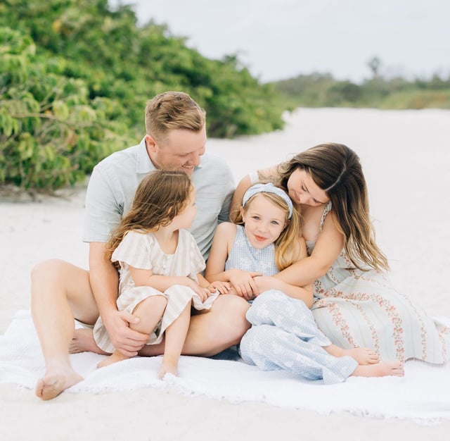 Family of four with two young girls sitting on a white blanket in the sand during a beach photo session