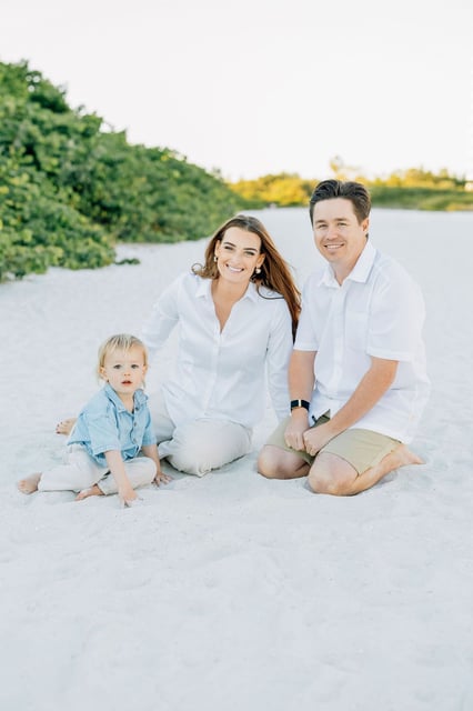 A family in white shirts sits smiling on white sand beside beach dune greenery at golden hour