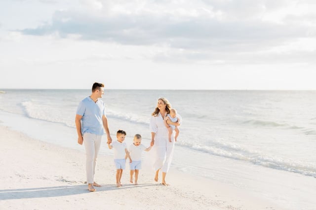 A family in white and light blue walks barefoot along the shoreline on a bright sandy Gulf beach