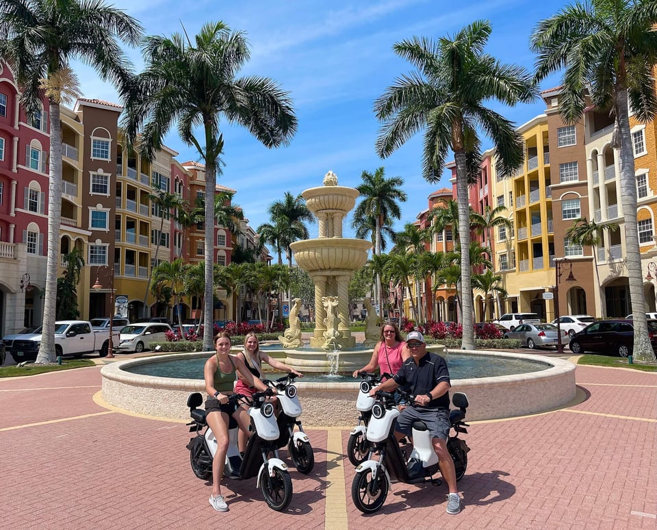 Four riders on electric mopeds pause by the tiered fountain at Bayfront Naples, framed by palms and pastel buildings