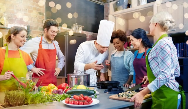 A chef in a white toque leads a group of smiling students in aprons around a kitchen counter piled with fresh vegetables