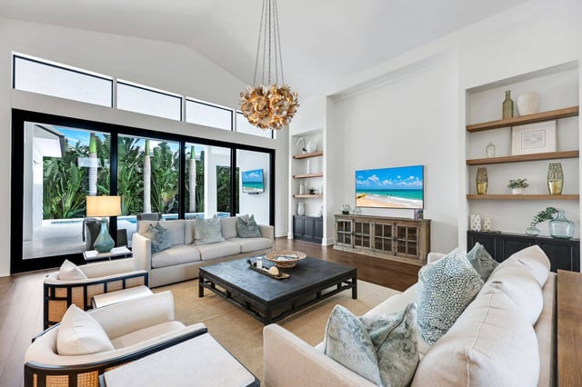 Bright vaulted living room with white sofas, gold pendant chandelier and sliding glass doors to a palm-lined pool