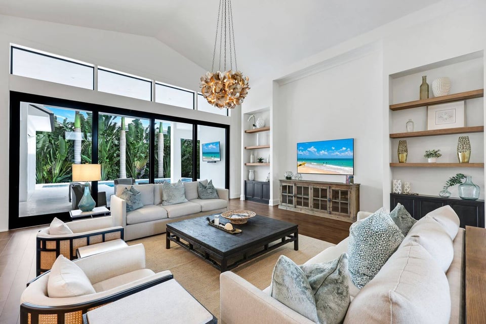 Bright vaulted living room with white sofas, gold pendant chandelier and sliding glass doors to a palm-lined pool