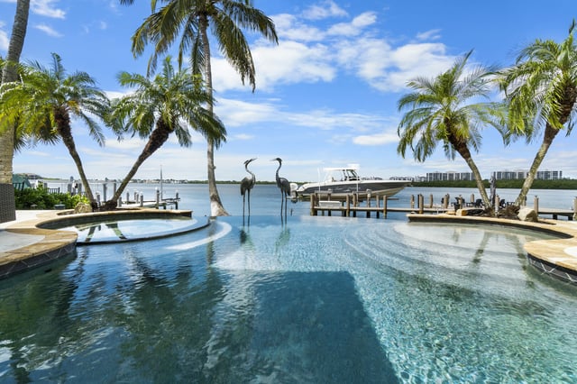 Infinity pool with heron sculptures overlooking a waterfront dock, boat lift and palm trees under blue sky