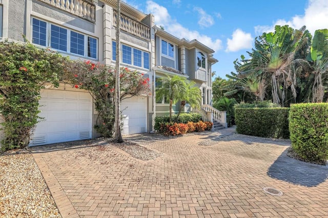 Brick paver driveway leading to a multi-story townhome with white garage doors, palms and tropical landscaping