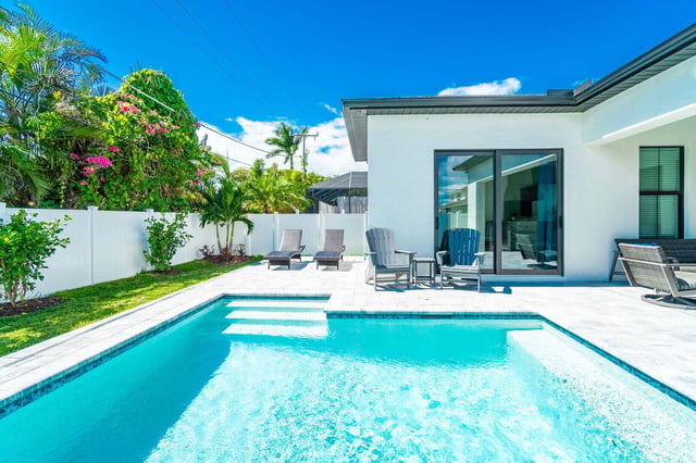 Backyard pool with sun shelf, lounge chairs and palms beside a white modern home under blue sky