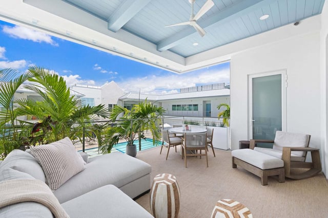 Covered outdoor terrace with pale blue beadboard ceiling, white sectional sofa and dining set overlooking a pool and palms
