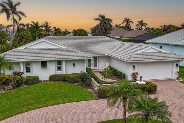 Aerial view of a single-story white ranch home with tile roof, paver driveway and palms at sunset
