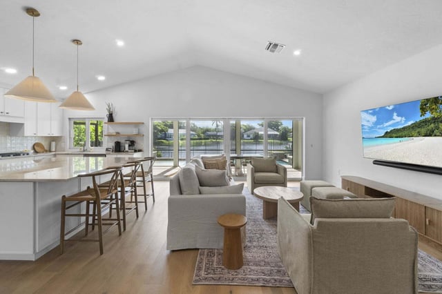 Open-concept living room with vaulted ceiling, linen sofas, kitchen island bar stools and sliding glass doors to a waterfront view