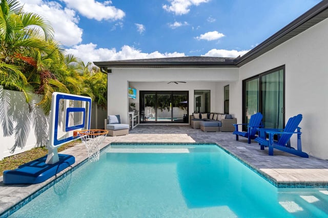 Backyard pool with a blue basketball hoop, Adirondack chairs and a covered patio under sunny skies