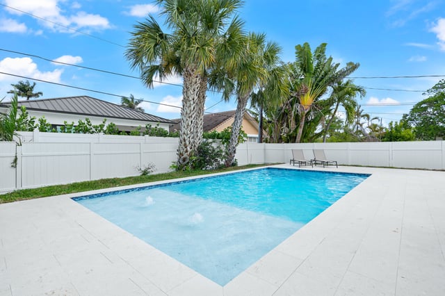 Backyard swimming pool with white deck, two lounge chairs, palm trees and a white vinyl privacy fence under blue sky