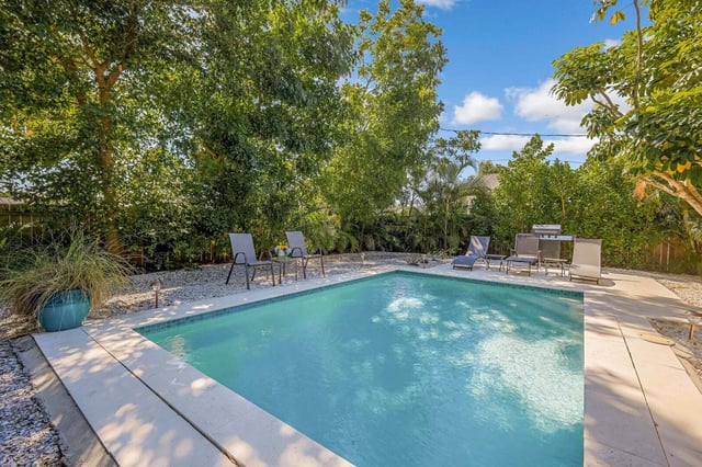 Backyard swimming pool surrounded by lounge chairs, gravel decking and lush tropical trees under a blue sky