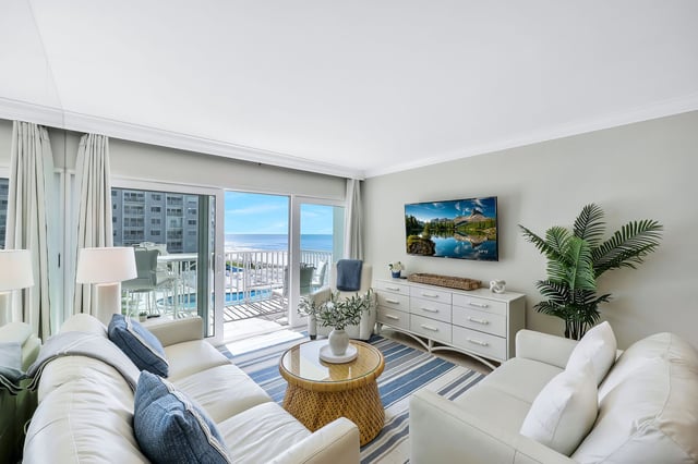 Bright coastal condo living room with white sofas, striped rug, wall-mounted TV and sliding doors to a Gulf-view balcony