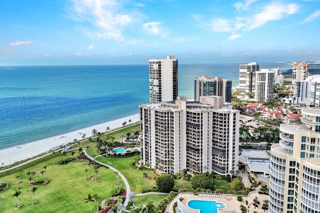 Aerial view of beachfront high-rise condominiums, palm-lined lawns and pools along turquoise Gulf waters in Naples