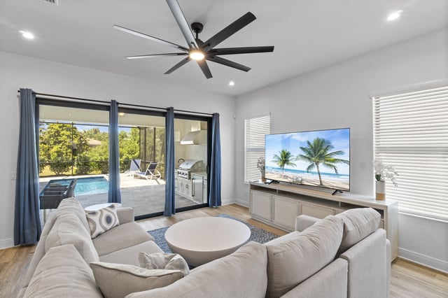 Bright living room with sectional sofa, ceiling fan and large TV, sliding doors opening to a screened pool lanai