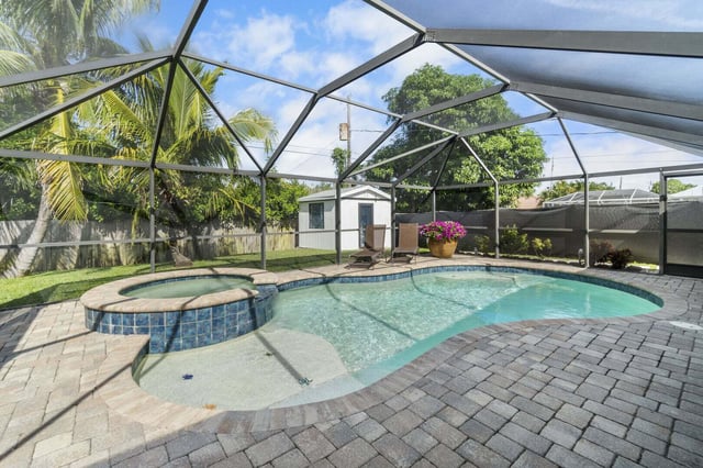 Screened lanai enclosing a kidney-shaped pool and attached spa with paver deck, palms and backyard shed beyond