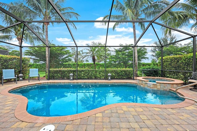 Screened lanai pool with attached spa, paver deck and lounge chairs framed by palms under a blue sky
