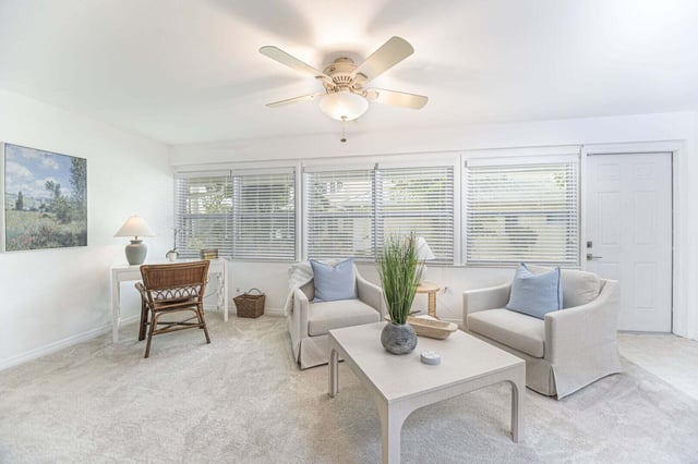 Bright living room with white slipcovered sofa and armchair, ceiling fan, blinds and pale carpet