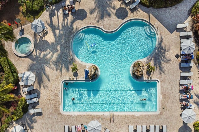 Aerial view of a curved resort pool with lap lanes, hot tub, white umbrellas and loungers on a paver deck