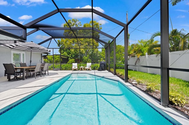 Screened lanai enclosing a turquoise pool with patio dining set, striped umbrella and palms under blue sky